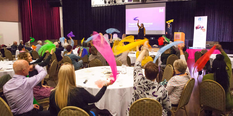 Delegates at round table, waving colourful scarves as part of a session on running engaging activities. In the background a speaker is on stage in front of a powerpoint presentation.