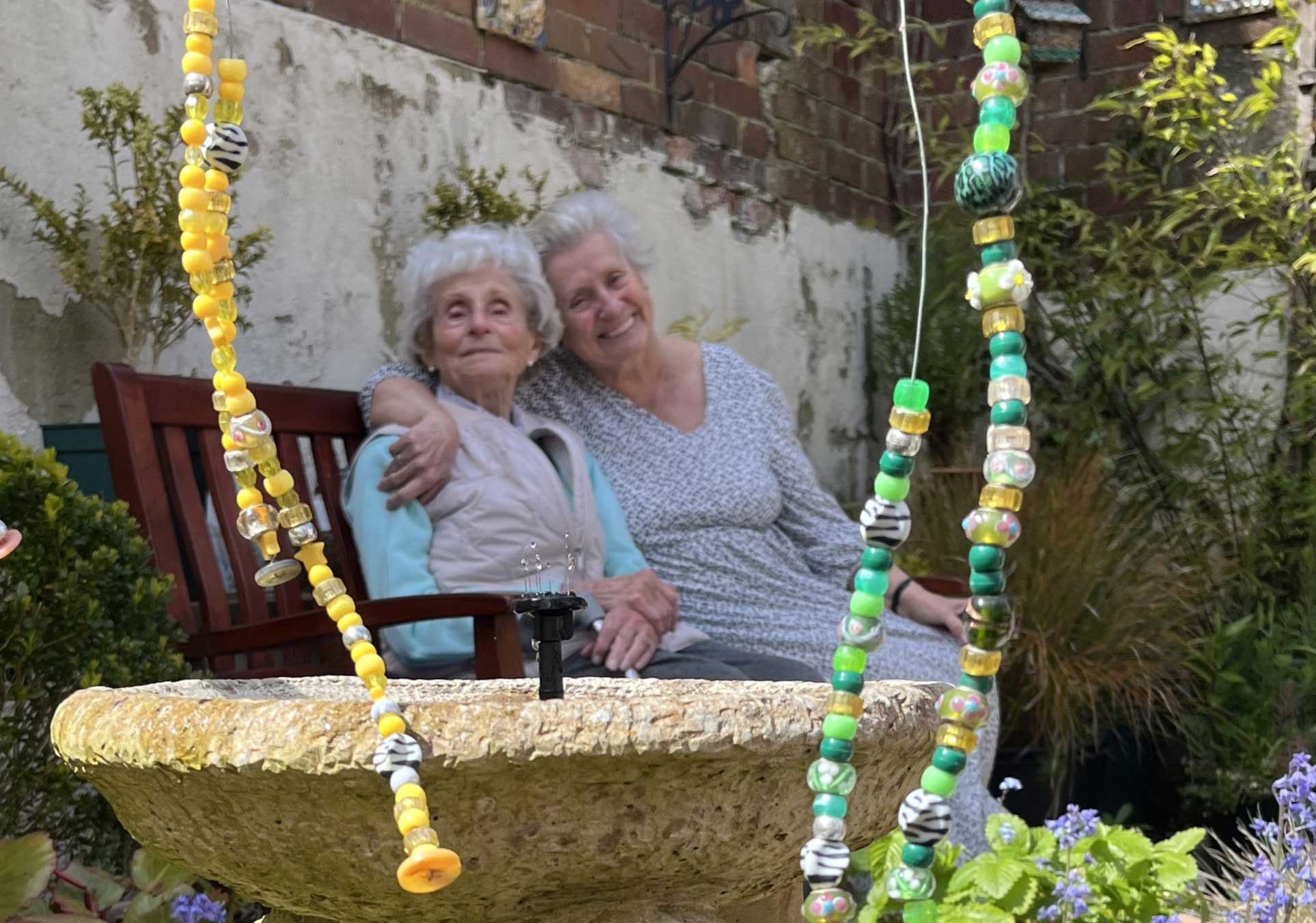 Two older females sitting together smiling in Sidmouth Street garden framed by coloured beads and a bird bath 
