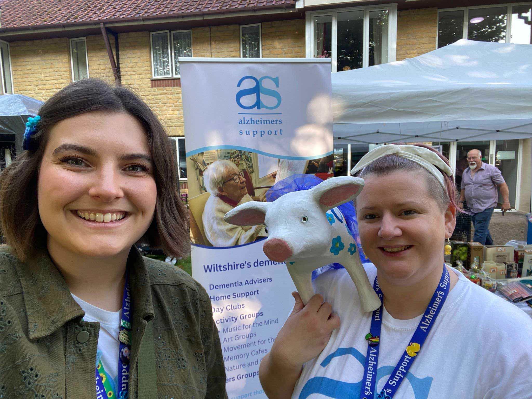 Two ladies, one in a dark jacket and one in an AS T shirt, holding a pottery pig with an Alzheimers Support banner and stall in the background 