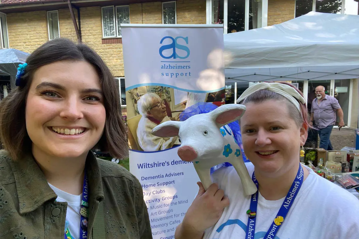 Two ladies, one in a dark jacket and one in an AS T shirt, holding a pottery pig with an Alzheimers Support banner and stall in the background