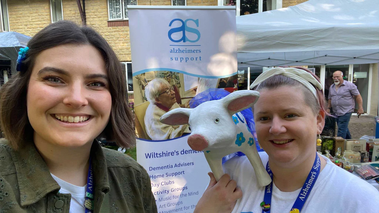 Two ladies, one in a dark jacket and one in an AS T shirt, holding a pottery pig with an Alzheimers Support banner and stall in the background