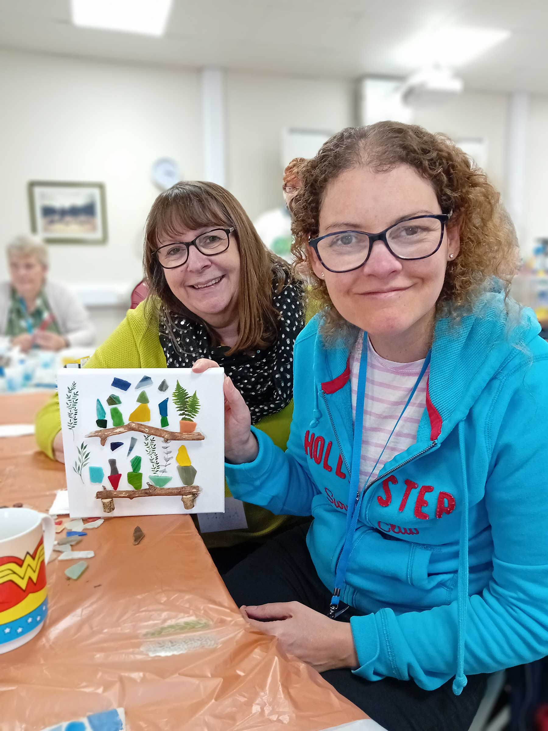 Two smiling ladies one with a blue hoody and one with a spotty scarf, holding a collage picture of two wooden shelves with pot plants on, taken at REME Muddy Boots