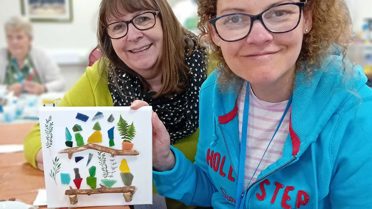 Two smiling ladies one with a blue hoody and one with a spotty scarf, holding a collage picture of two wooden shelves with pot plants on, taken at REME Muddy Boots