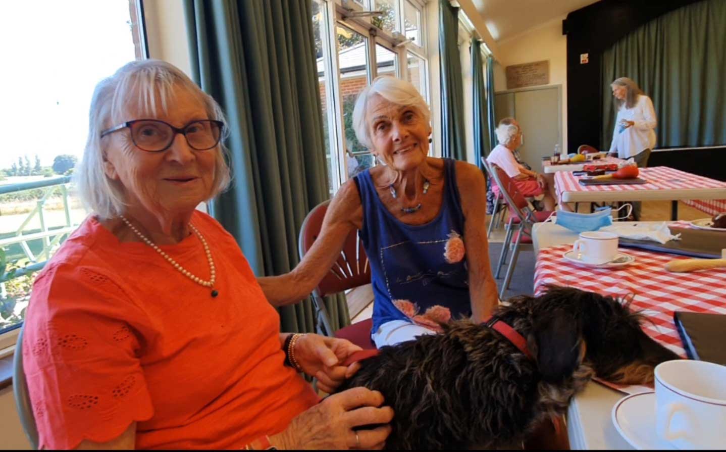 A lady with an orange T shirt and necklace sitting with a dog on her lap resting his head on the table.  Another lady in a blue vest to sits next to them at Lockeridge Music Group 