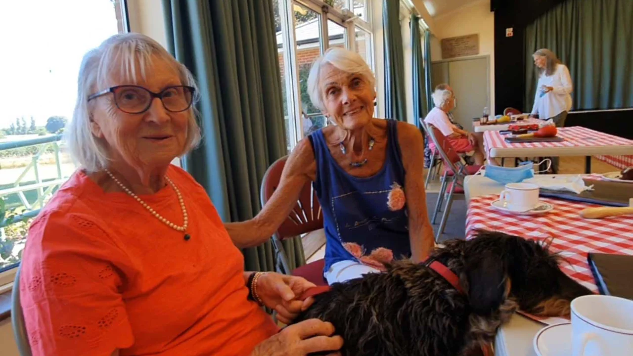A lady with an orange T shirt and necklace sitting with a dog on her lap resting his head on the table. Another lady in a blue vest to sits next to them at Lockeridge Music Group