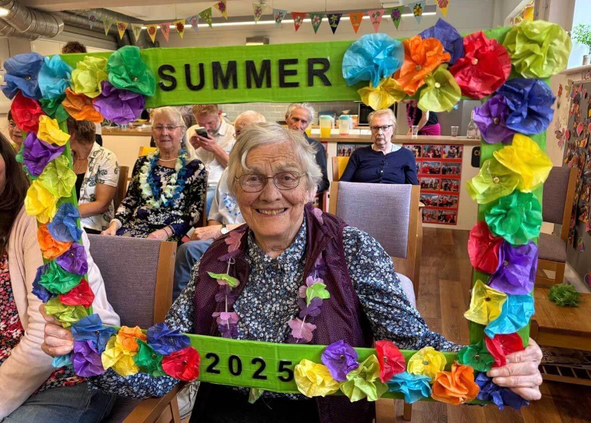 A lady in blue smiling through a flower coloured frame saying Summer 2025 with people sitting in the background at Scots Lane Club 