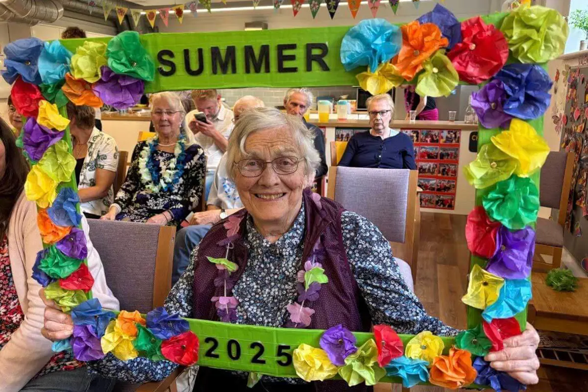 A lady in blue smiling through a flower coloured frame saying Summer 2025 with people sitting in the background at Scots Lane Club