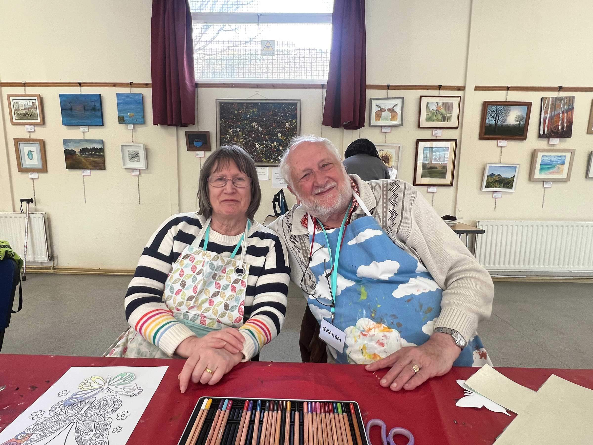 Beneficiaries Graham and Alwyn leaning in together as they take part in an art activity. Both are smiling and wearing colourful cloth aprons 