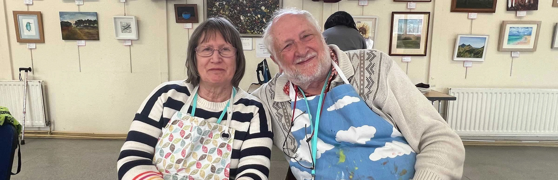 Beneficiaries Graham and Alwyn leaning in together as they take part in an art activity. Both are smiling and wearing colourful cloth aprons Beneficiaries Graham and Alwyn leaning in together as they take part in an art activity. Both are smiling and wearing colourful cloth aprons