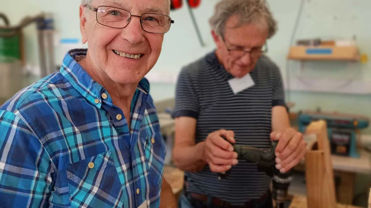 Smiling man in a checked shirt standing next to a work bench with man next to him who is drilling into a piece of wood at Corsham Men's Shed