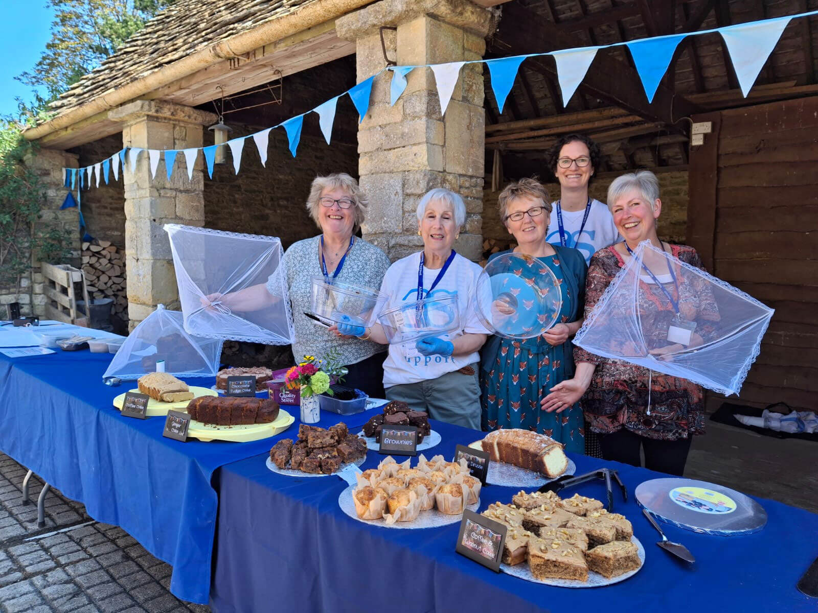 Group of lovely smiling ladies on a sunny day in front of a stone barn with blue bunting, standing behind a cake table with eight different cakes and blue table cloths.  