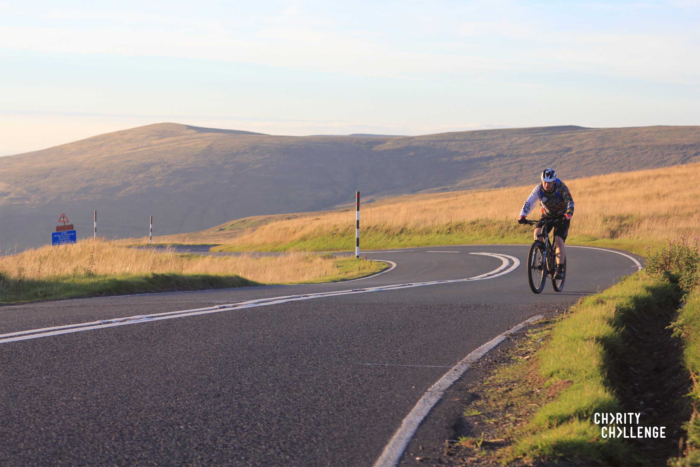 A beautiful scene of a lone cyclist riding near the top of a hill with moorland grass and a hill in the background.  Charity Challenge is written in the right hand corner