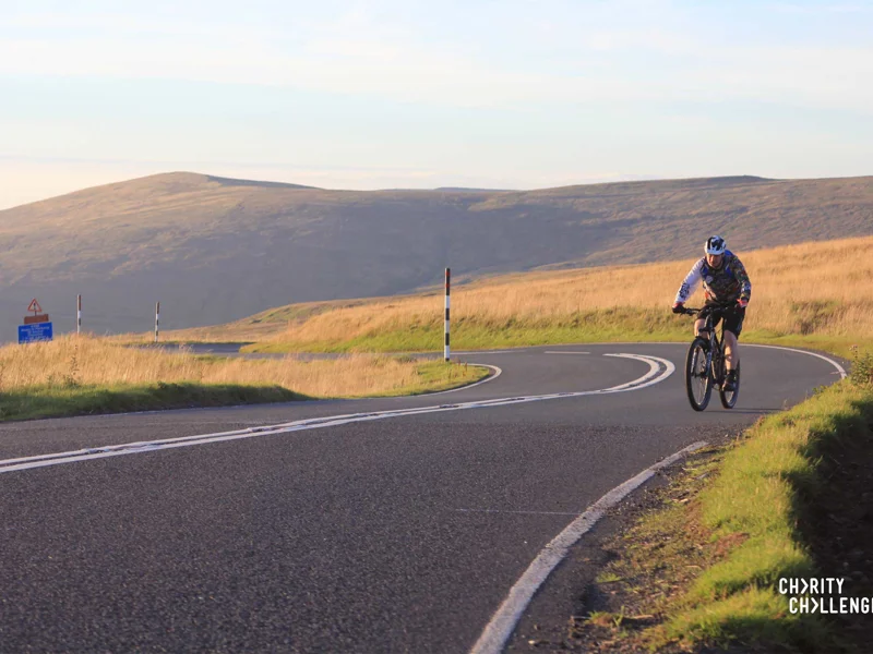 A beautiful scene of a lone cyclist riding near the top of a hill with moorland grass and a hill in the background.  Charity Challenge is written in the right hand corner A beautiful scene of a lone cyclist riding near the top of a hill with moorland grass and a hill in the background.  Charity Challenge is written in the right hand corner