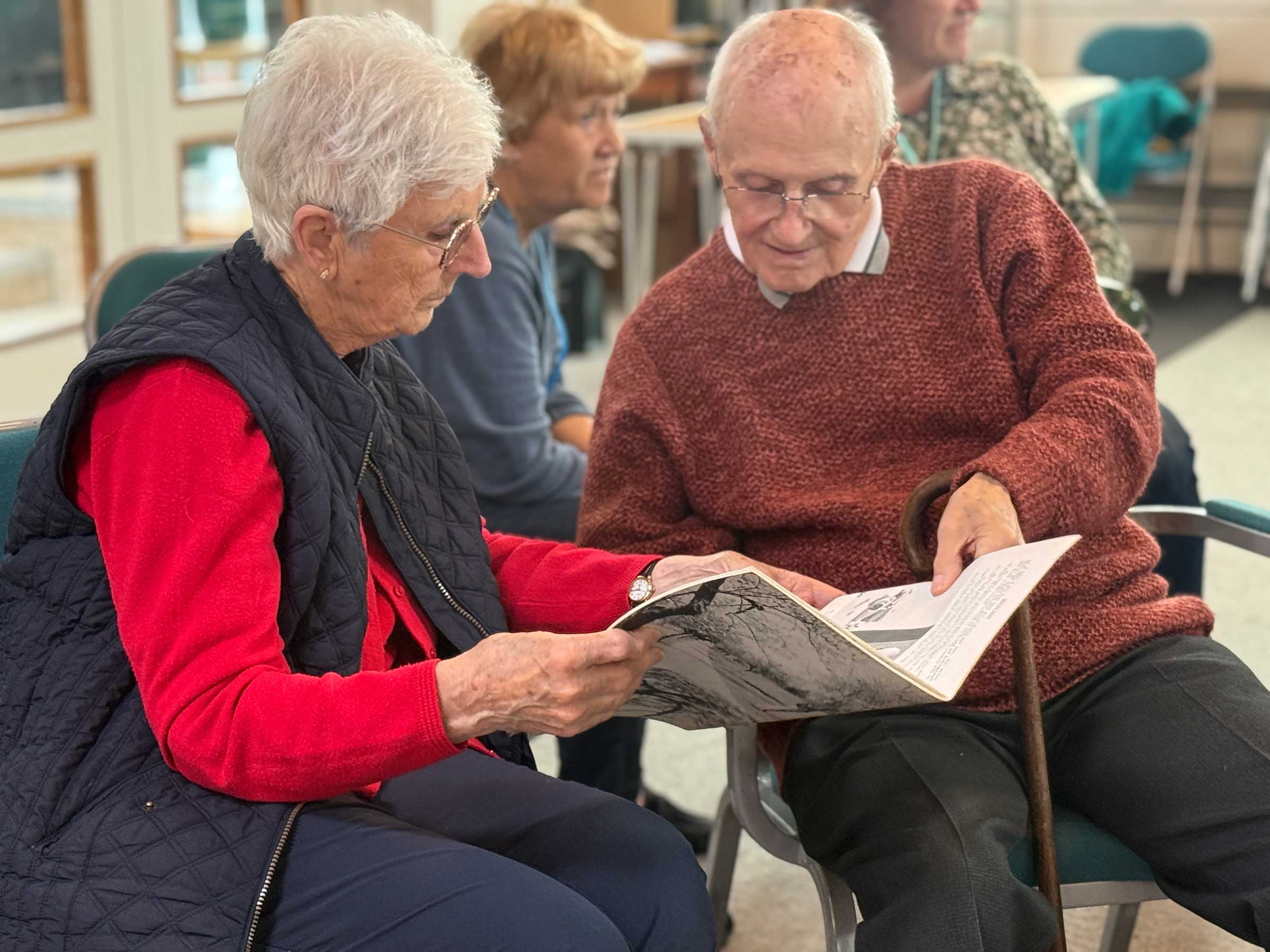 A man and woman seated together at a memory cafe, engrossed in an old booklet they are both holding. Wearing jumpers in autumn colours
