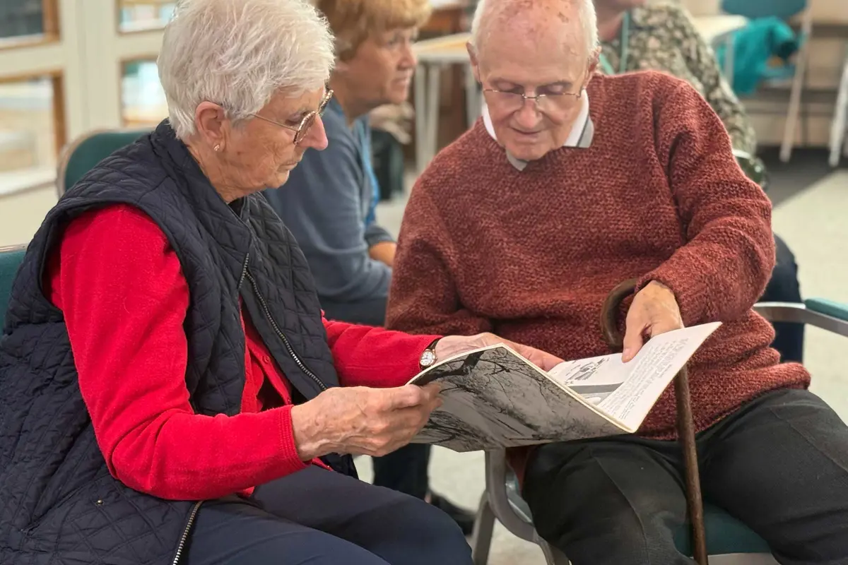 A man and woman seated together at a memory cafe, engrossed in an old booklet they are both holding. Wearing jumpers in autumn colours