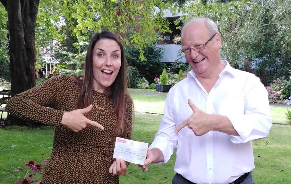 Fundraiser Laura and supporter Alan pulling happy faces as they point to a normal sized cheque in front of a tree in a park