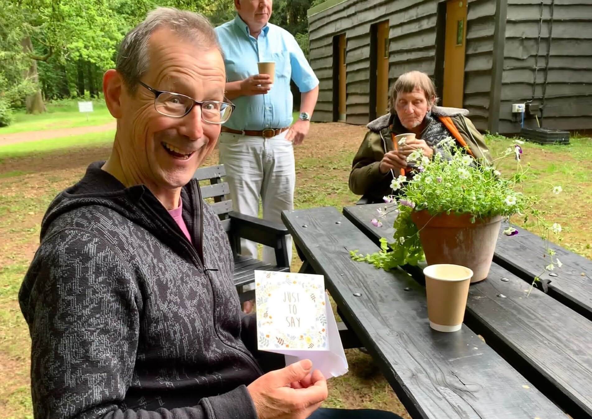 A gentleman volunteer in dark top sits at a picnic table facing us and holding a cards which says Just to Say.  In the background there is a man in blue shirt standing next to a man seated with a coat around his shoulders with trees and a brown building