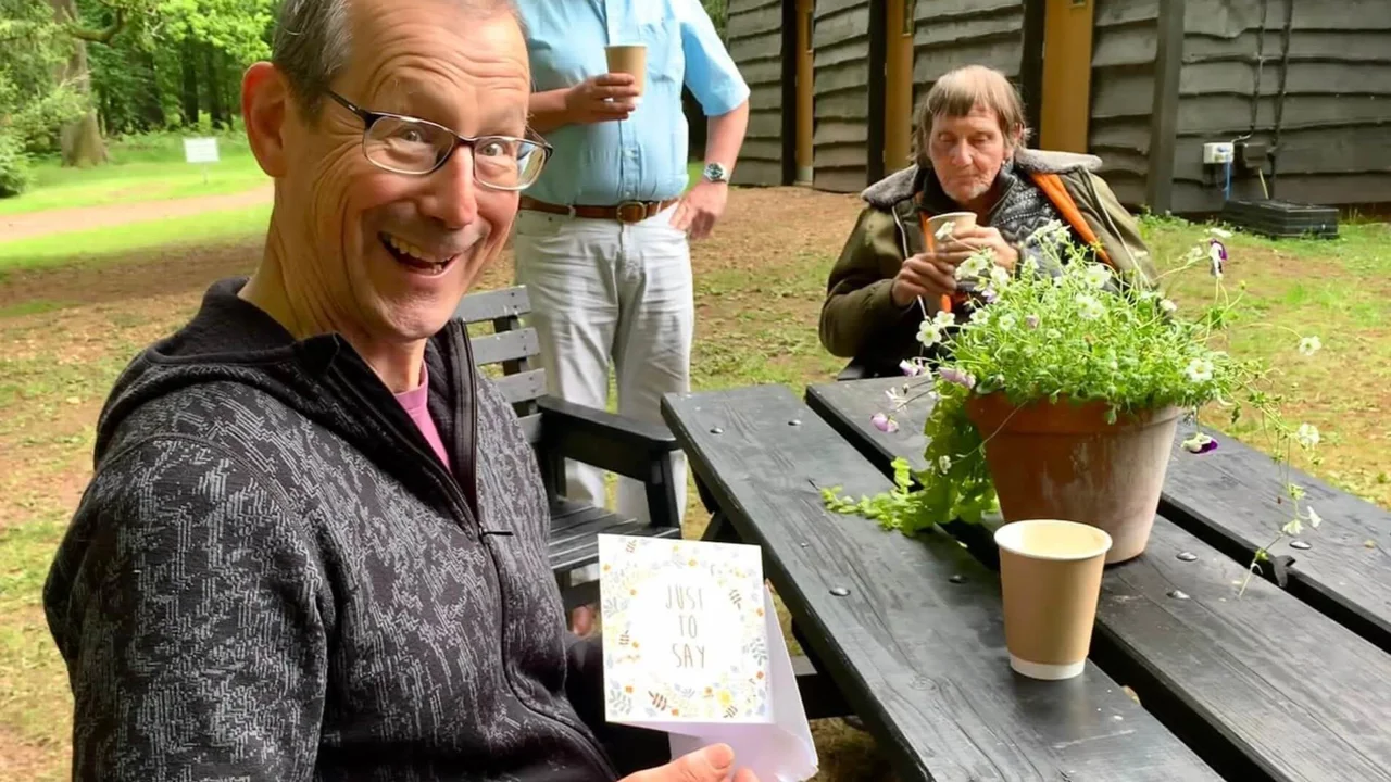 A gentleman volunteer in dark top sits at a picnic table facing us and holding a cards which says Just to Say. In the background there is a man in blue shirt standing next to a man seated with a coat around his shoulders with trees and a brown building