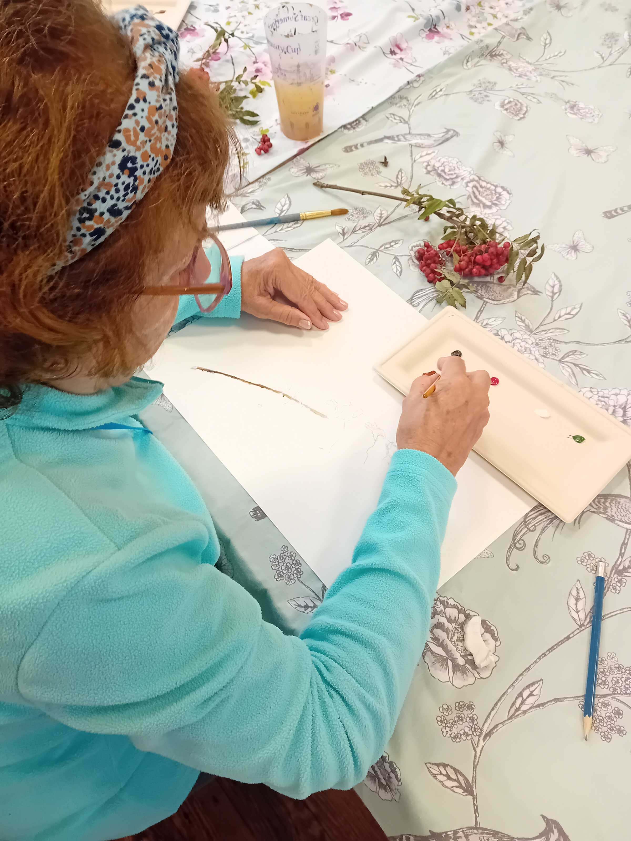 Close up picture of a ladies with a headband and blue top looking down on a piece of paper, her hand is holding a paint brush and there is a paint tray and the life object of a green branch with red berries on it.  Taken at Lyneham Art group 