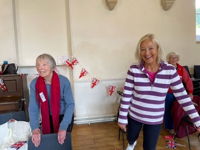 An older lady stands behind her chair with her hands on it, the other lady in a striped top is standing with one foot raised behind her, both are smiling and there is union jack bunting behind them