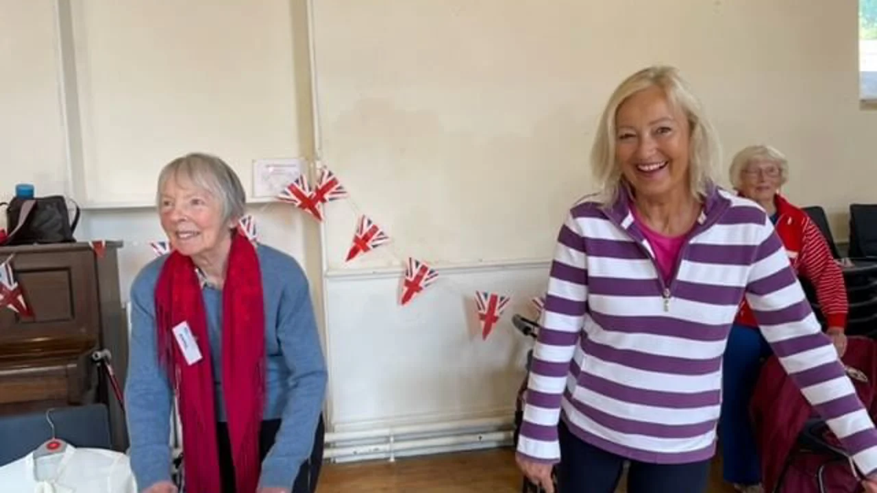 An older lady stands behind her chair with her hands on it, the other lady in a striped top is standing with one foot raised behind her, both are smiling and there is union jack bunting behind them