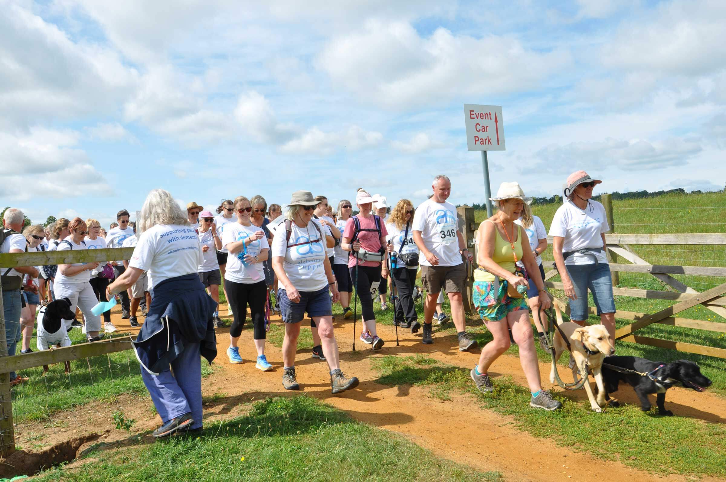 A crowd of walkers starting out together through a wide gate. It's a blue-sky day and most are wearing shorts and charity branded white T-shirts
