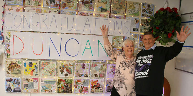 Duncan and Lyn Baxter waving in front of a Congratulations Duncan banner at Old Silk Works club