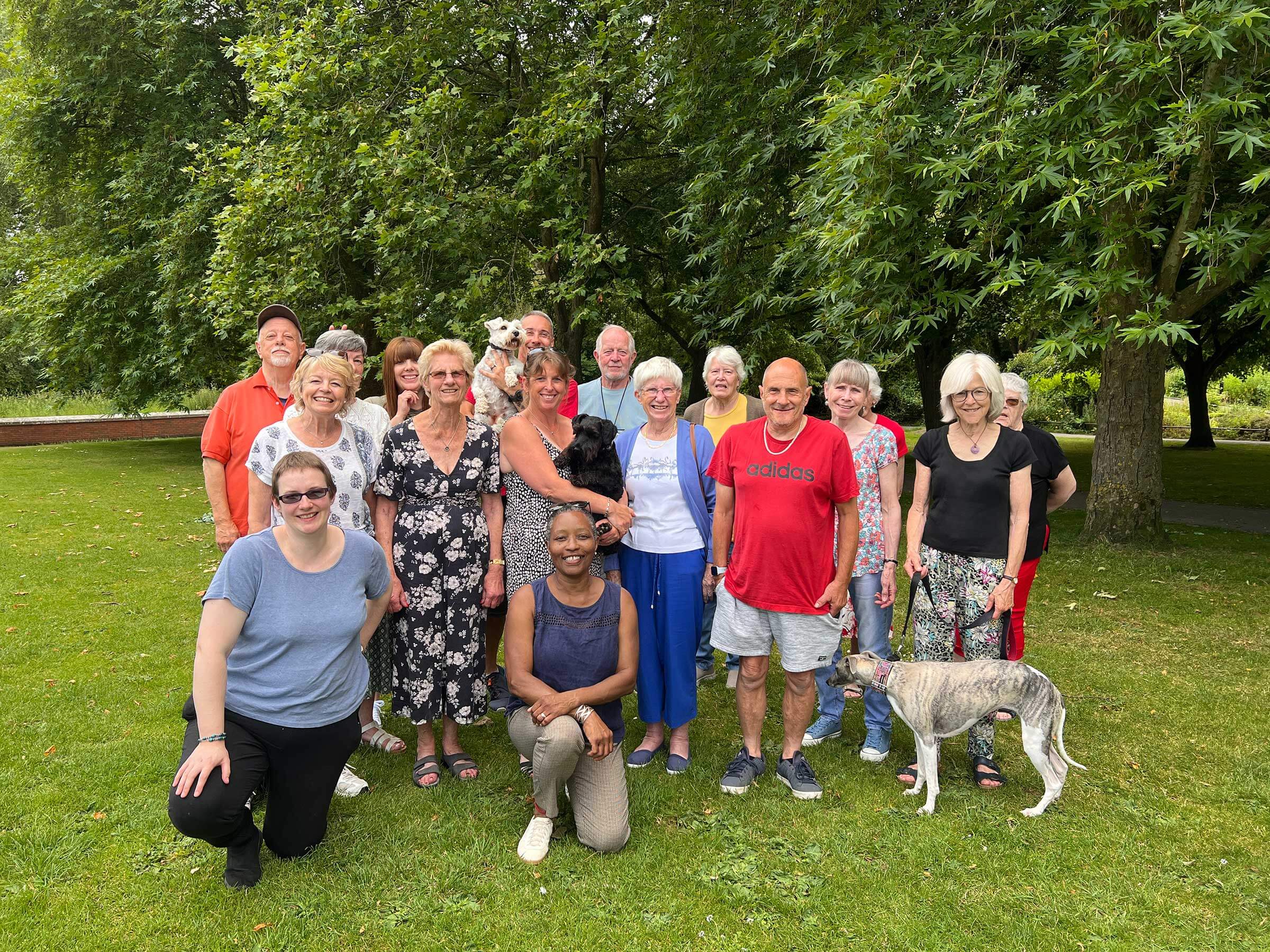 A group of 16 smiling volunteers and three dogs standing together on the grass in front of trees in a park