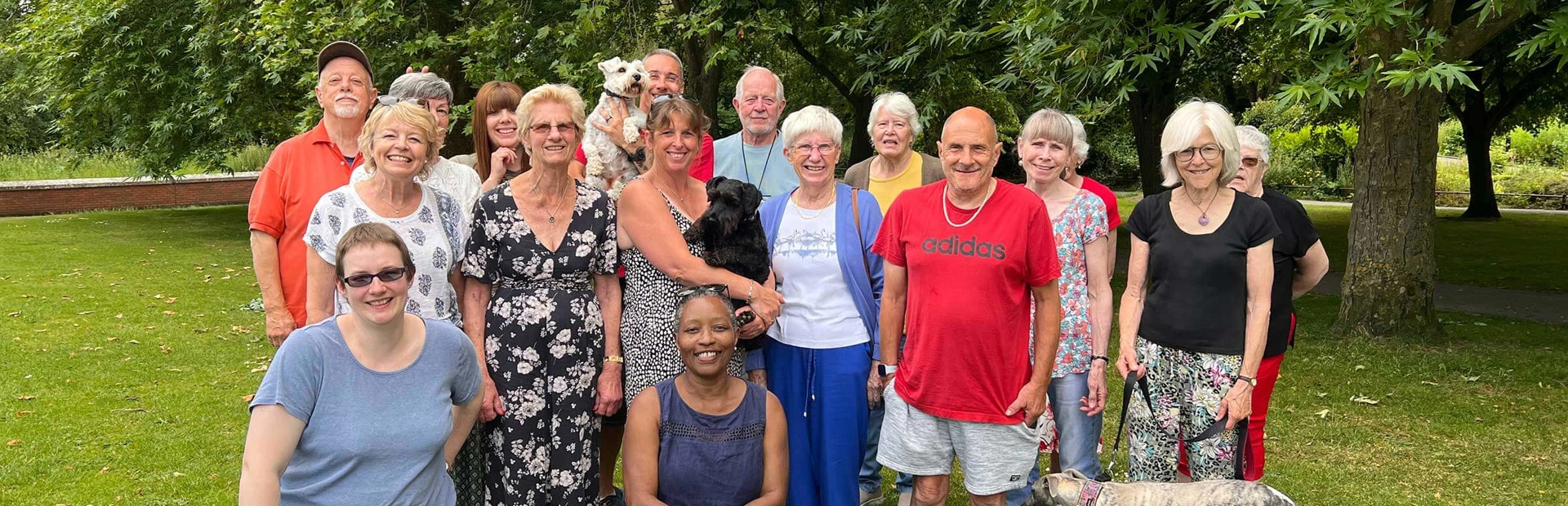 A group of 16 smiling volunteers and three dogs standing together on the grass in front of trees in a park A group of 16 smiling volunteers and three dogs standing together on the grass in front of trees in a park