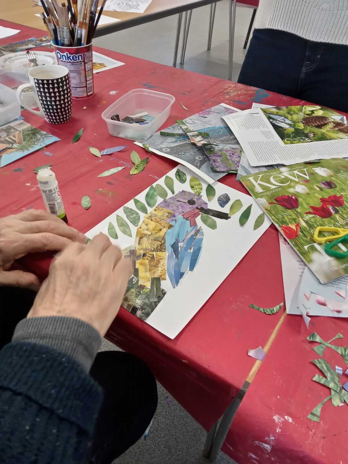 Close up picture of a collaged bird with blue, yellow and purple flowers.  Around the picture is magazines, cut out pictures, a pot, a cup and a glue stick, all taken at Warminster Art group 