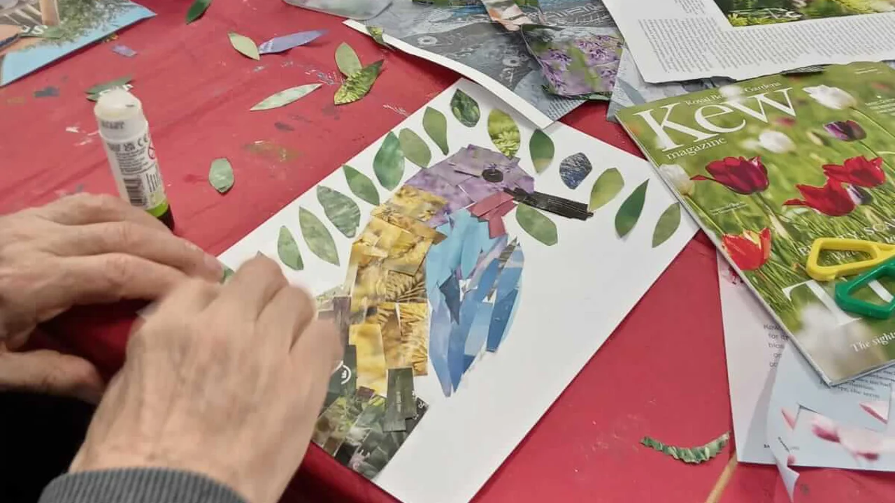 Close up picture of a collaged bird with blue, yellow and purple flowers. Around the picture is magazines, cut out pictures, a pot, a cup and a glue stick, all taken at Warminster Art group