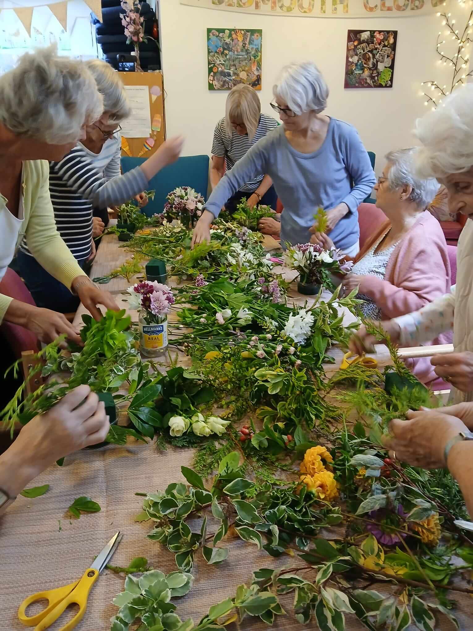 Group of women in Sidmouth Street Club choosing flowers from a table covered in greenery and pink, white and yellow flowers 