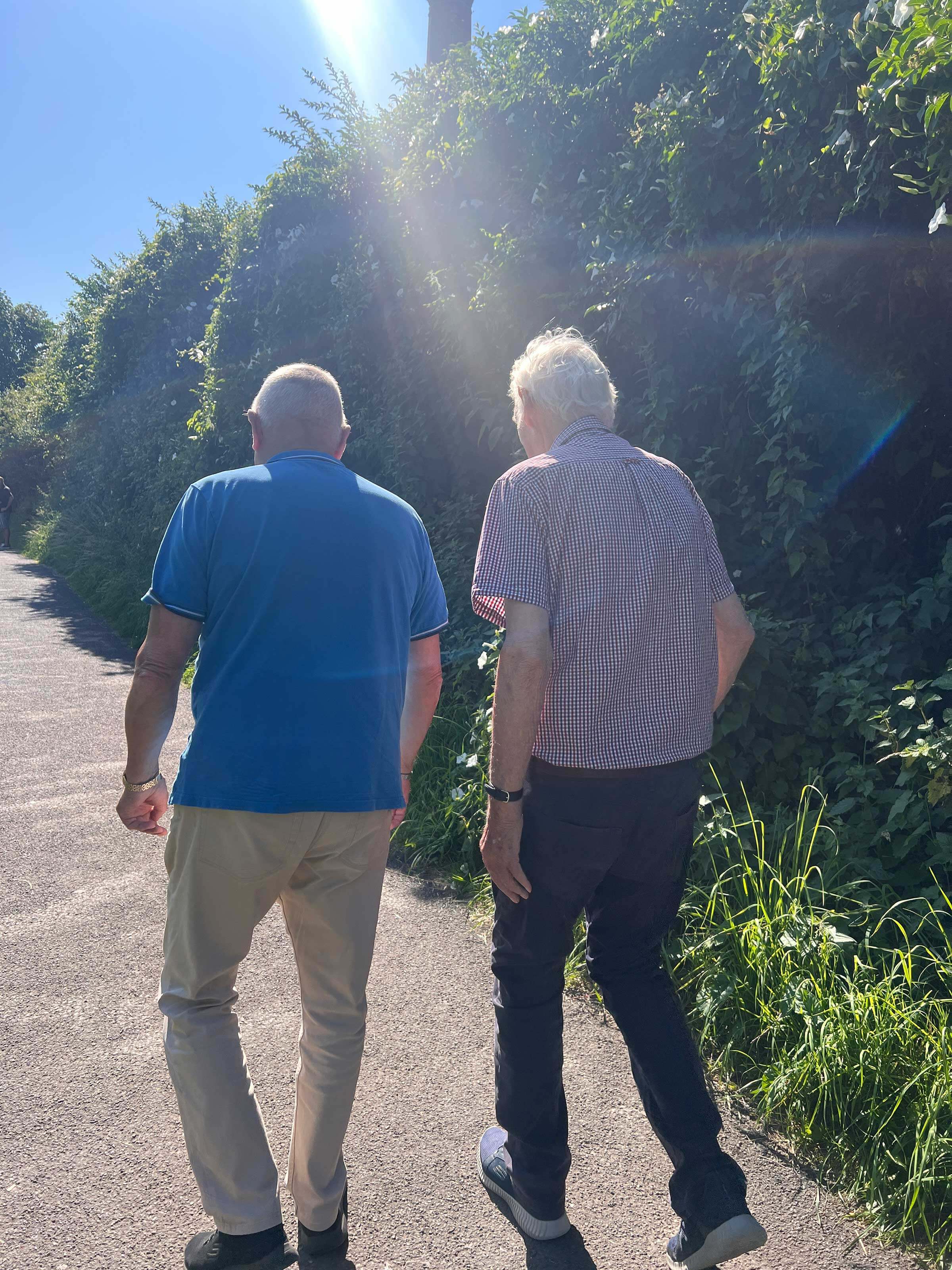 Two older gentlemen in short sleeve shirts walk away from the camera down a gravel path on a beautiful sunny day