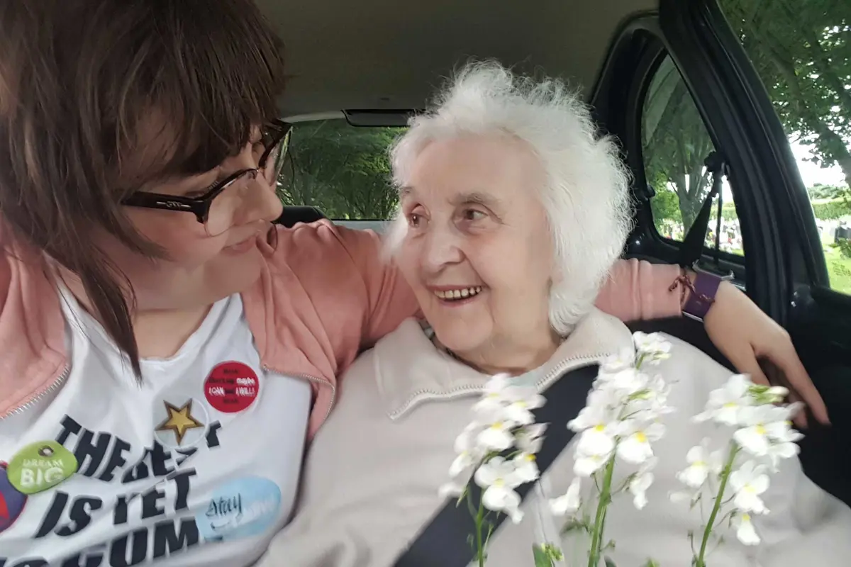 This is a close up loving picture of two ladies sitting in a car together, one has her arm round the other, they are smiling at each other,  The older lady is holding a plant which has white flowers on it.