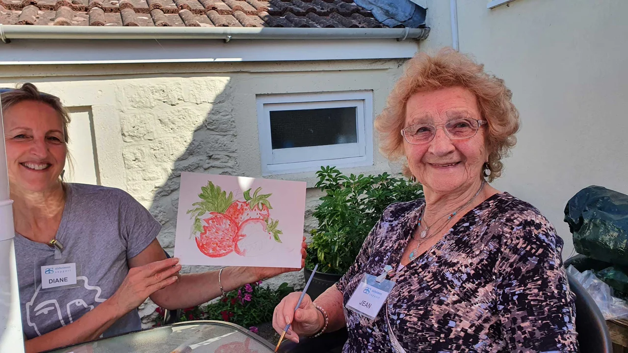 Two ladies are sitting in the shade of an umbrella. The younger lady holds up a picture of strawberries painted in red and green. The older lady holds a paint brush and is smiling. Taken at Lyneham Art Group