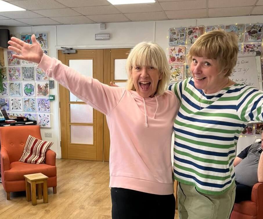 Two volunteers standing and waving in Old Silk Works club - one has a pink top and the other a stripy blue and green top with arm chairs and picture behind them 
