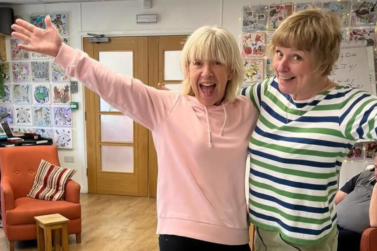 Two volunteers standing and waving in Old Silk Works club - one has a pink top and the other a stripy blue and green top with arm chairs and picture behind them