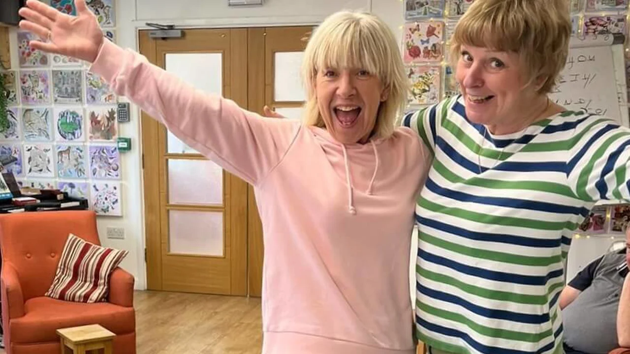 Two volunteers standing and waving in Old Silk Works club - one has a pink top and the other a stripy blue and green top with arm chairs and picture behind them