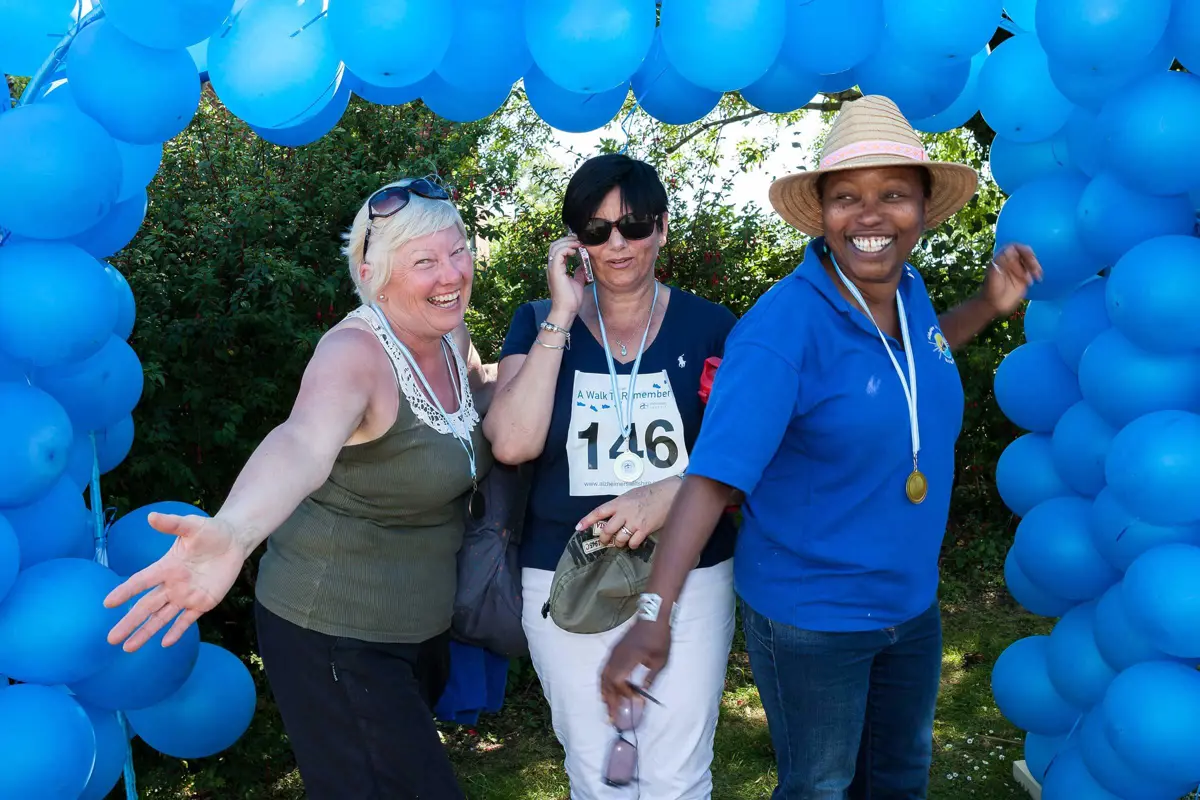 Three ladies with Walk to Remember medals on standing beneath a blue balloon arch smiling, laughing and with their arms outstretched in celebration