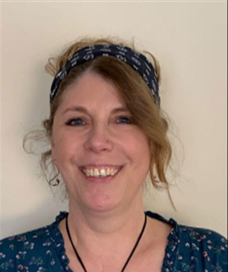 headshot of a smiling Sue with a hair band holding back her wavy hair, in a blue print top