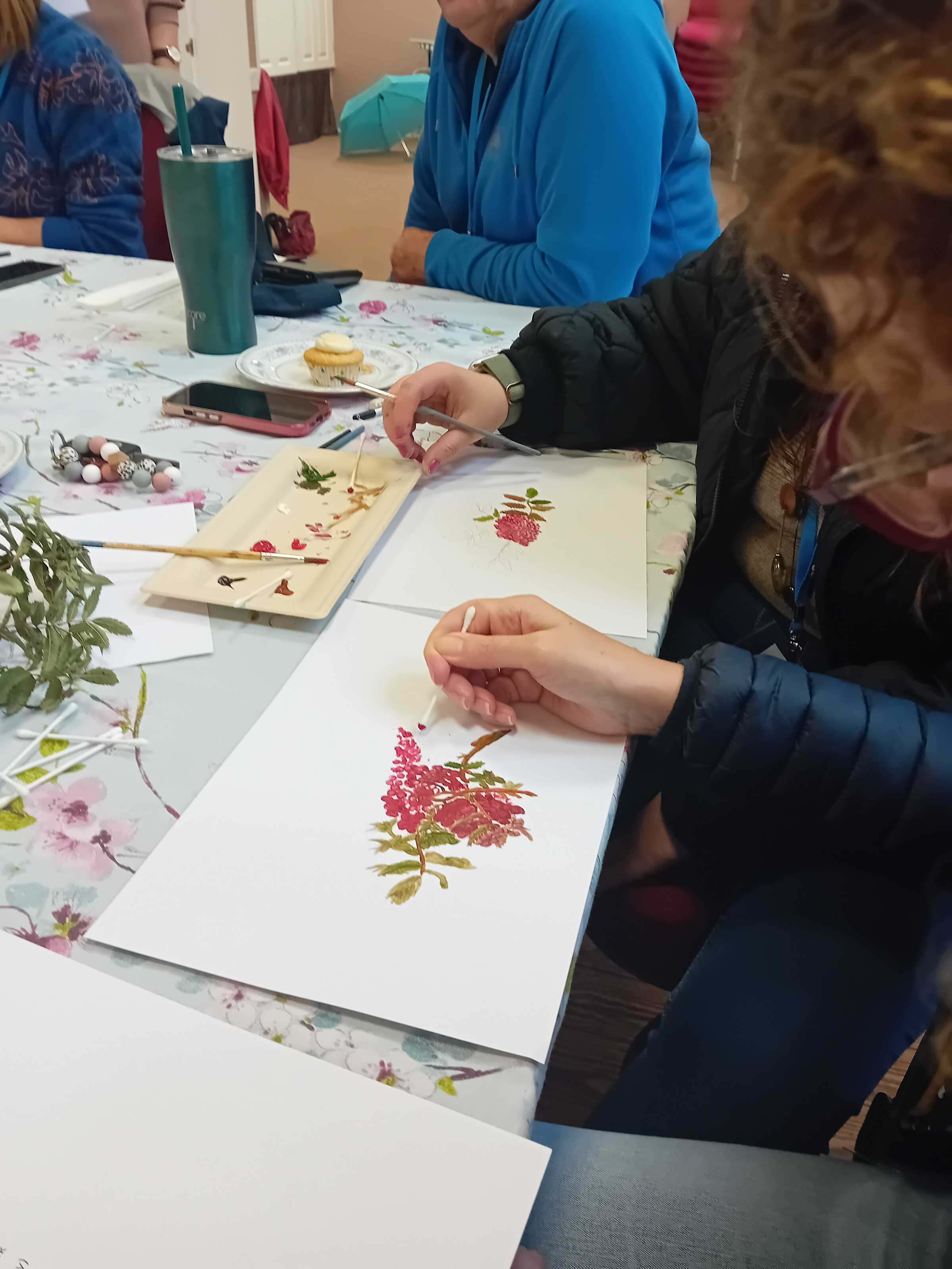 Close up picture of a ladies hands whilst she paints a picture of a flowered stem in pink and green using an earbud.  Around her is a paint dish, a plant, earbuds, another hand of someone painting and a phone.  All taken at Lyneham Art group  