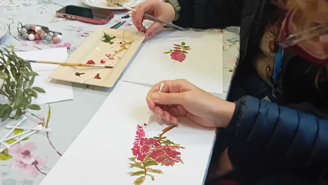Close up picture of a ladies hands whilst she paints a picture of a flowered stem in pink and green using an earbud. Around her is a paint dish, a plant, earbuds, another hand of someone painting and a phone. All taken at Lyneham Art group