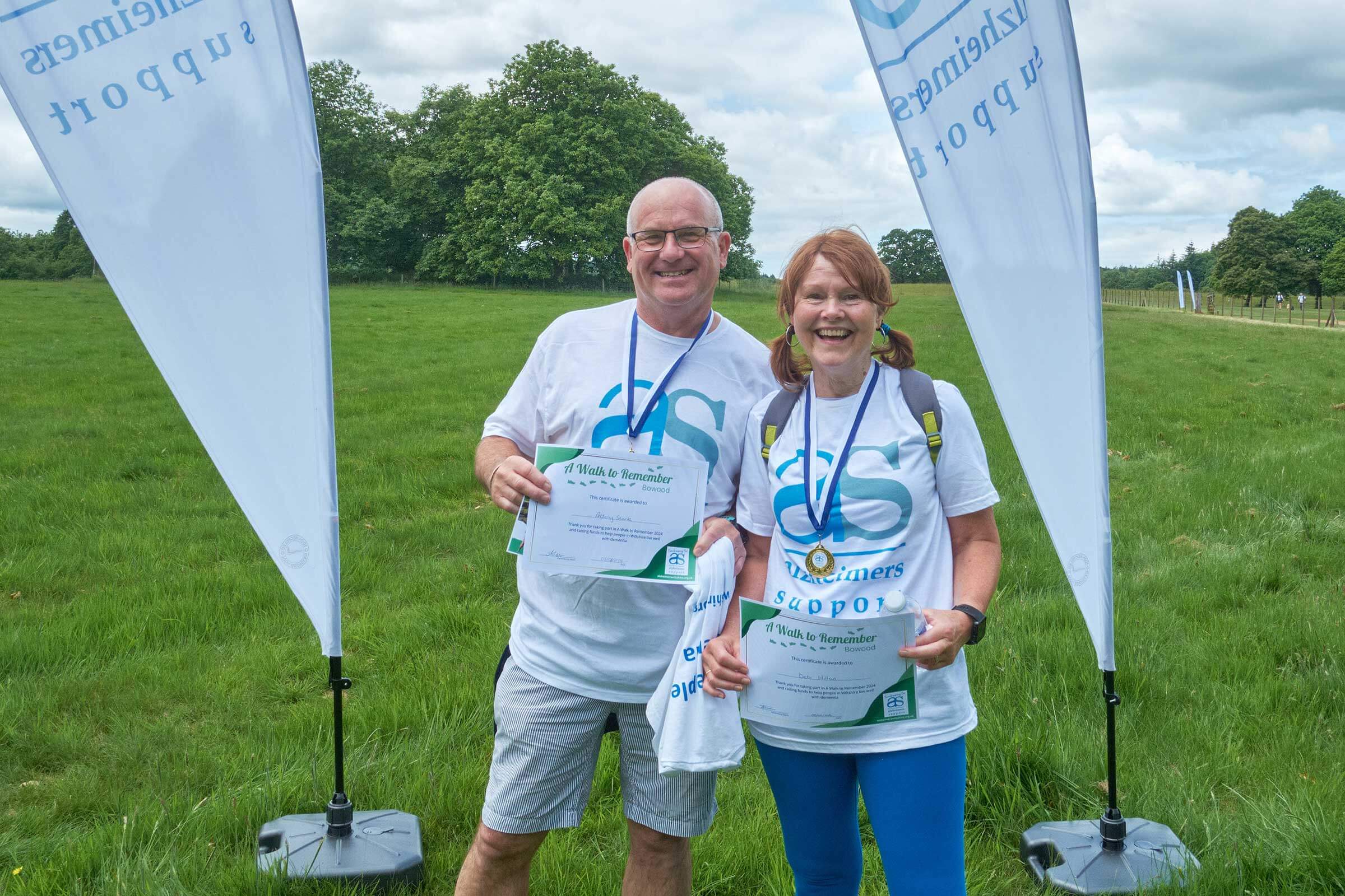 A man and a woman wearing Alzheimer Support t shirts, wearing medals and holding certificates and standing in a field between two sail flags at Bowood having just completed A walk to Remember 