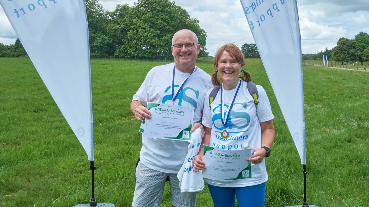 A man and a woman wearing Alzheimer Support t shirts, wearing medals and holding certificates and standing in a field between two sail flags at Bowood having just completed A walk to Remember