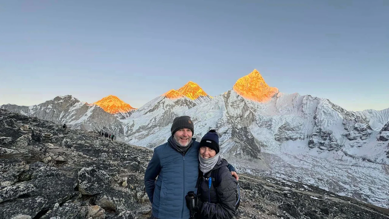 A man in a bobble hat, blue jacket and scarf with a lady in a black coat, scarf and bobble hat are standing in a rocky mountainous setting with sun shining on the peaks, one of them Everest behind them.