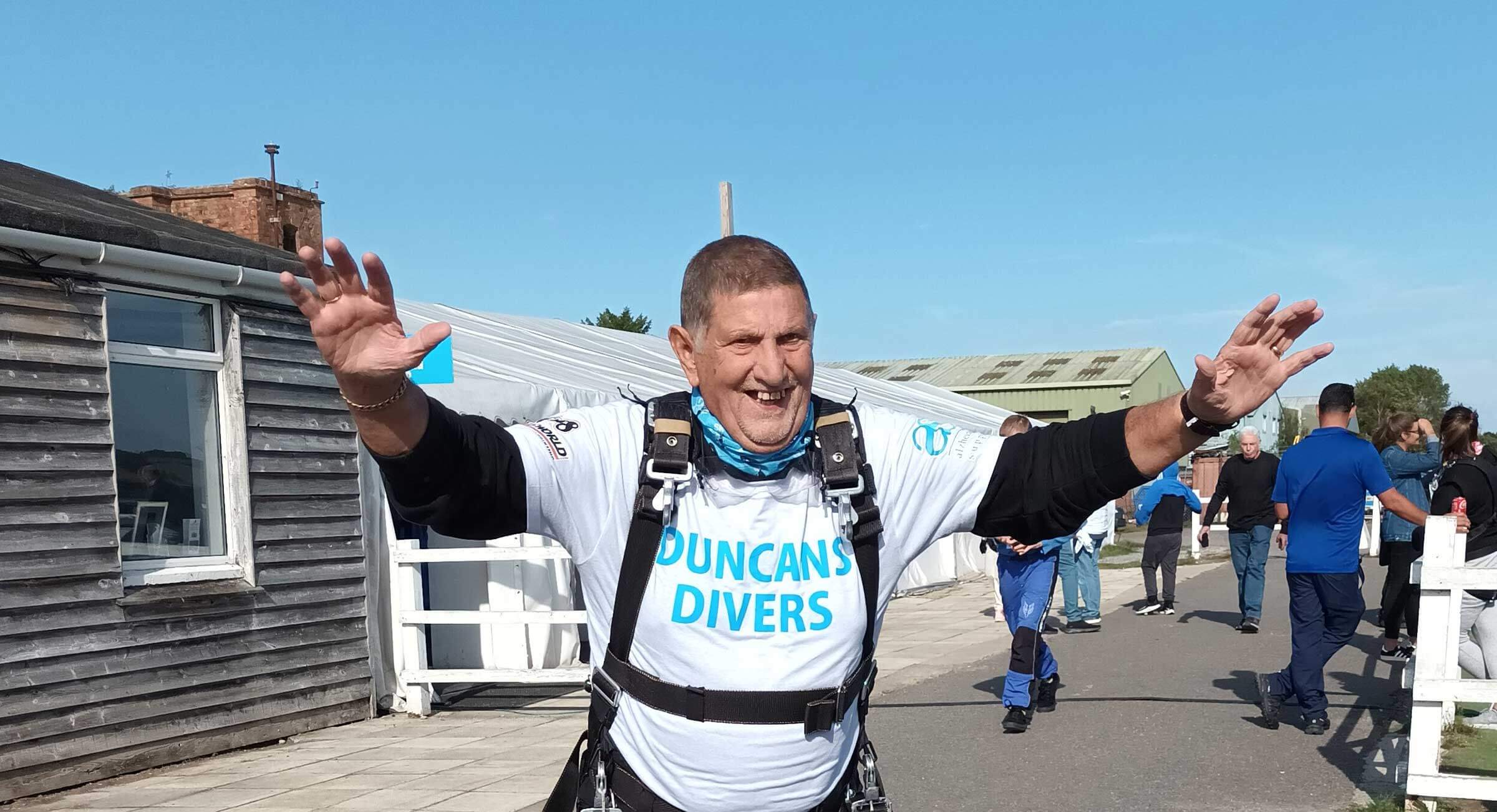 Duncan, wearing a sky dive harness, white T Shirt and neck warmer standing with the arms raised to celebrate he has doing a Sky Dive 