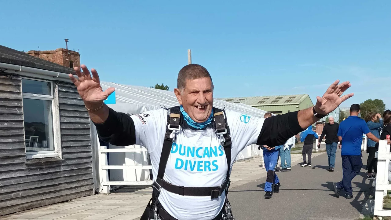 Duncan, wearing a sky dive harness, white T Shirt and neck warmer standing with the arms raised to celebrate he has doing a Sky Dive