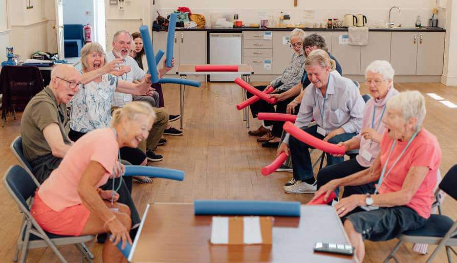 A group of men and women moving and playing a game with red and blue foam tubes, they are laughing and looking forwards at Chippenham Movement for the Mind 