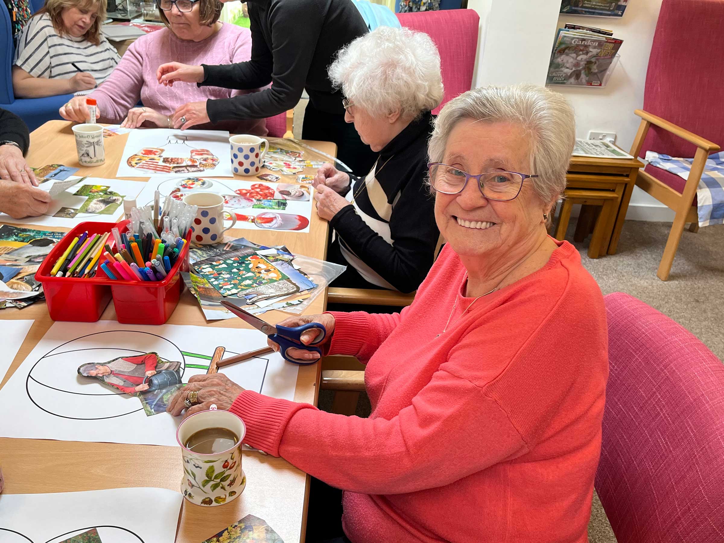A smiling lady wearing a coral jumper sitting with a group of people all drinking cups of tea and collaging at Sidmouth Street 