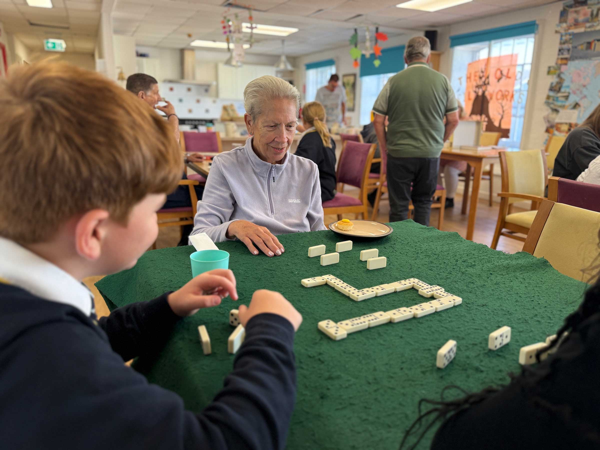 A schoolboy plays dominoes with a female club member on a green baize table with other club activities in the background
